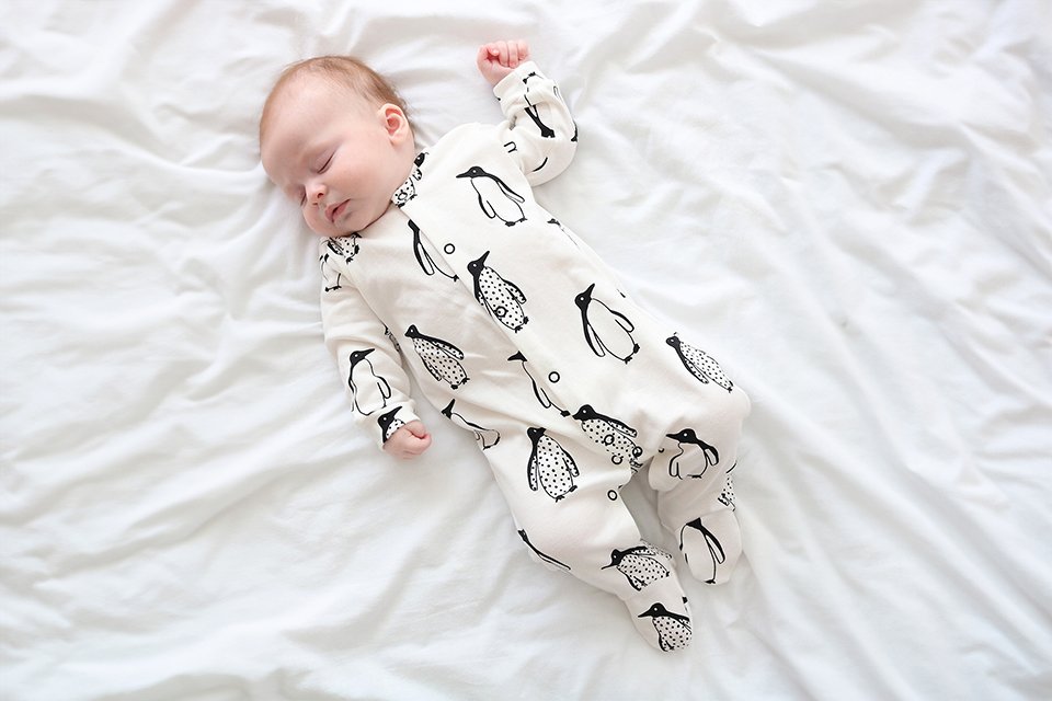 A baby in a FRED & NOAH Milk Penguin sleepsuit sleeping on the bed.